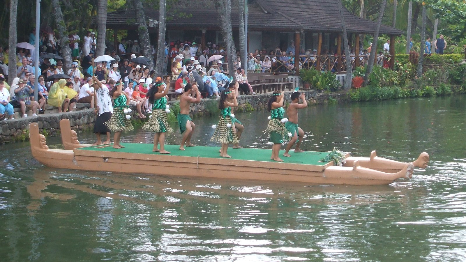 Dancers on a long canoe on the Polynesian Cultural Center lagoon with crowds watching