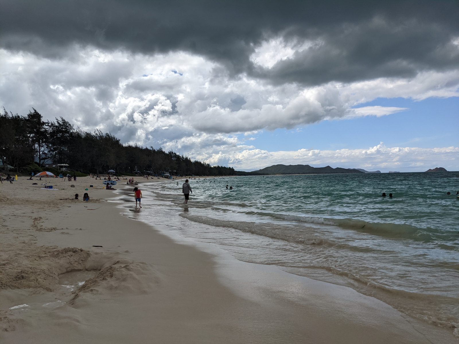 Waimanalo Beach shoreline — beachgoers and swimmers along the long sandy bay, ironwood trees behind the sand, May 2021